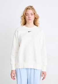 Sudadera blanca hecha de un material suave con cuello redondo, hombros caídos y un pequeño logo negro de Nike centrado en el pecho.