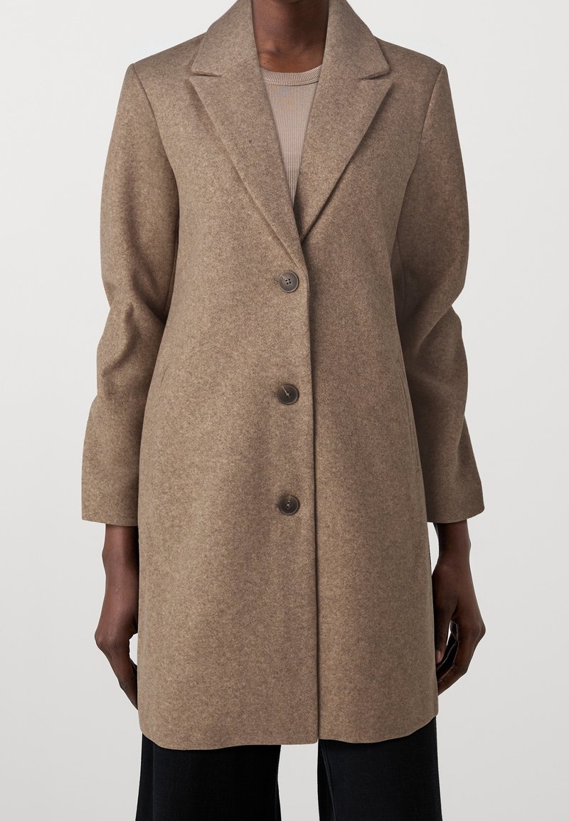 Manteau en laine marron longueur genoux avec trois boutons, porté sur un top côtelé beige et un pantalon noir.