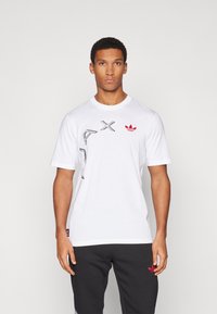 White cotton t-shirt featuring graphic prints in black and a red Adidas logo on the chest. Short sleeves and round neckline.