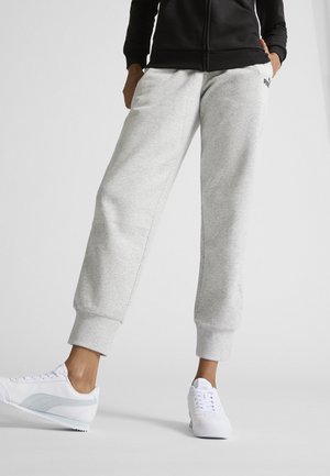 Personne portant un pantalon de survêtement gris clair, une veste noire zippée, des baskets blanches, debout avec une main dans la poche.