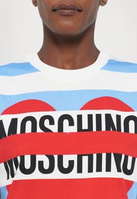 Crewneck t-skjorte med blå, hvite og røde horisontale striper. "Moschino" trykket med fet svarte bokstaver over et rødt hjerte-design.