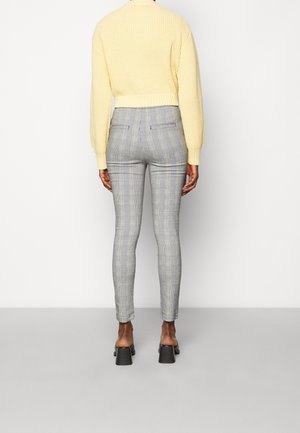 Persona con pantalones ajustados de cuadros grises, sandalias negras de tacón y un jersey de punto amarillo de manga larga, de pie junto a una pared blanca lisa.
