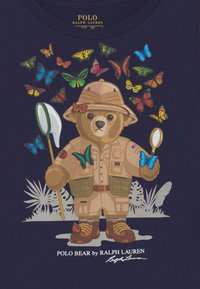 Tričko s kresleným medvědem v khaki oblečení, obklopeným barevnými motýly. Tmavě námořnické pozadí s detailním grafickým designem.