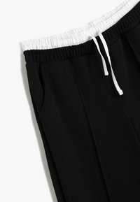 Zwarte broek met een witte elastische tailleband en trekkoord. Beschikt over zijzakken en een gladde, gestructureerde stof.