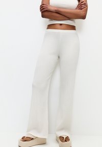 Femme portant un pantalon blanc à taille haute et large en côtelé avec des tongs plateformes crème, se tenant les bras croisés contre un mur blanc.