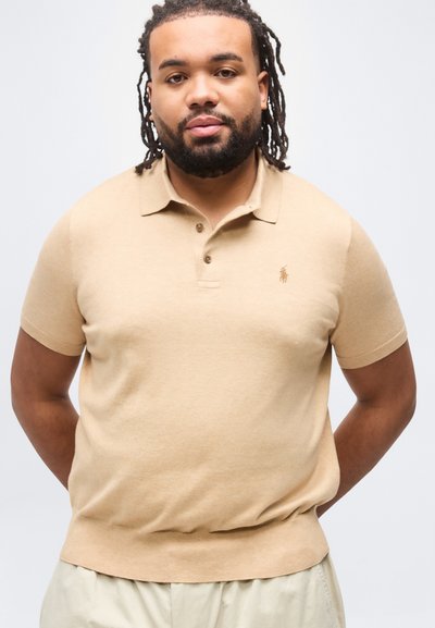 Άνδρας με πλεξούδες στα μαλλιά και γενειάδα, φοράει μπεζ κοντομάνικη πόλο μπλούζα και ανοιχτόχρωμο παντελόνι, στέκεται με τα χέρια πίσω από την πλάτη σε λευκό φόντο.