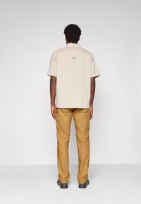 Homme de dos portant une chemise à manches courtes beige clair, un pantalon cargo moutarde et des chaussures noires sur un fond gris uni.
