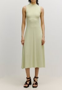 Ärmelloses knöchellanges Kleid aus hellgrünem, geripptem Stoff mit hohem Kragen und tailliertem Oberteil, das sich ab der Taille ausstellt. Braune, sandalartige High-Heels.