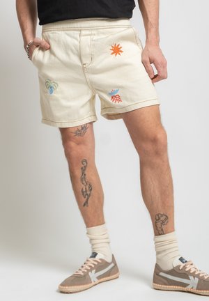 Man wearing cream shorts with colorful sun, crab, and plant embroidery, paired with beige sneakers and white socks, standing with hands in pockets.