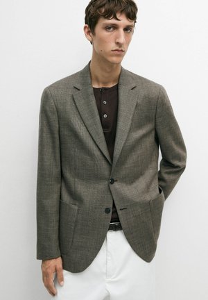 Giovane uomo che indossa un blazer grigio a quadri sopra una maglia henley scura e pantaloni bianchi, in piedi contro uno sfondo chiaro e semplice.