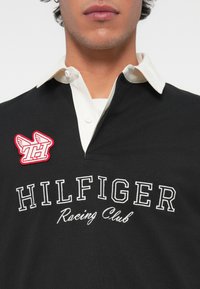 Melns polo krekls ar baltu apkakli, kas rotāts ar tekstu "HILFIGER Racing Club" un sarkanu izšūtu logo ar spārniem kreisajā pusē.