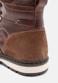 Tacco di stivale in pelle marrone e suede con cuciture bianche e suola in gomma bianca e nera su sfondo bianco.