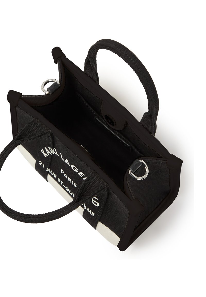 Sac à main en toile noire avec des accents blancs, double poignées, quincaillerie en argent et texte imprimé sur un côté. Forme rectangulaire compacte avec une ouverture sur le dessus.