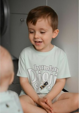 Vaaleanvihreä lyhythihainen t-paita, jossa on "humlan Djoj" -painatus ja mehiläiskuvasto. Lapsi istuu harmaalla lattialla, päällään vaaleat shortsit.