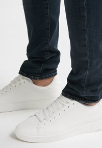 Persona vestida con jeans azul oscuro y zapatillas blancas de lona con cordones en una superficie blanca, mostrando solo las piernas inferiores y los pies.