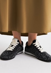 Zapatillas negras adornadas con brillantes plateados, con una suela blanca, cordones negros y un logo cerca del tobillo.