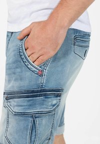 Denim shorts in light blue with a faded texture. Features pockets with contrasting stitching and a small red tag. Casual design.