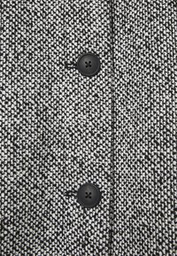 Abrigo de tela texturizada en blanco y negro con un patrón tejido, que presenta dos botones negros para el cierre.