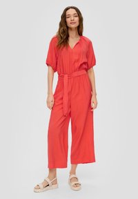 Koraalkleurige jumpsuit met korte mouwen, v-hals en een strikceintuur in de taille; heeft brede pijpen en een gestructureerde stof. G gedragen met chunky sandalen.