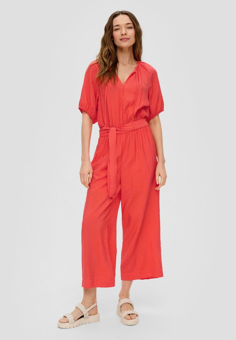 Koraalkleurige jumpsuit met korte mouwen, v-hals en een strikceintuur in de taille; heeft brede pijpen en een gestructureerde stof. G gedragen met chunky sandalen.