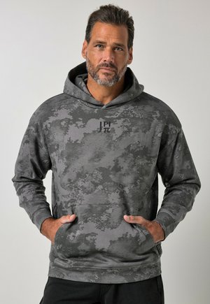 Hombre con barba corta que lleva una sudadera con capucha gris de estampado de camuflaje, con bolsillo delantero y pequeño logo negro en el pecho sobre fondo gris claro.