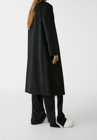 Largo abrigo negro de tela texturizada, combinado con pantalones negros holgados con franjas laterales blancas y zapatillas gruesas blancas.