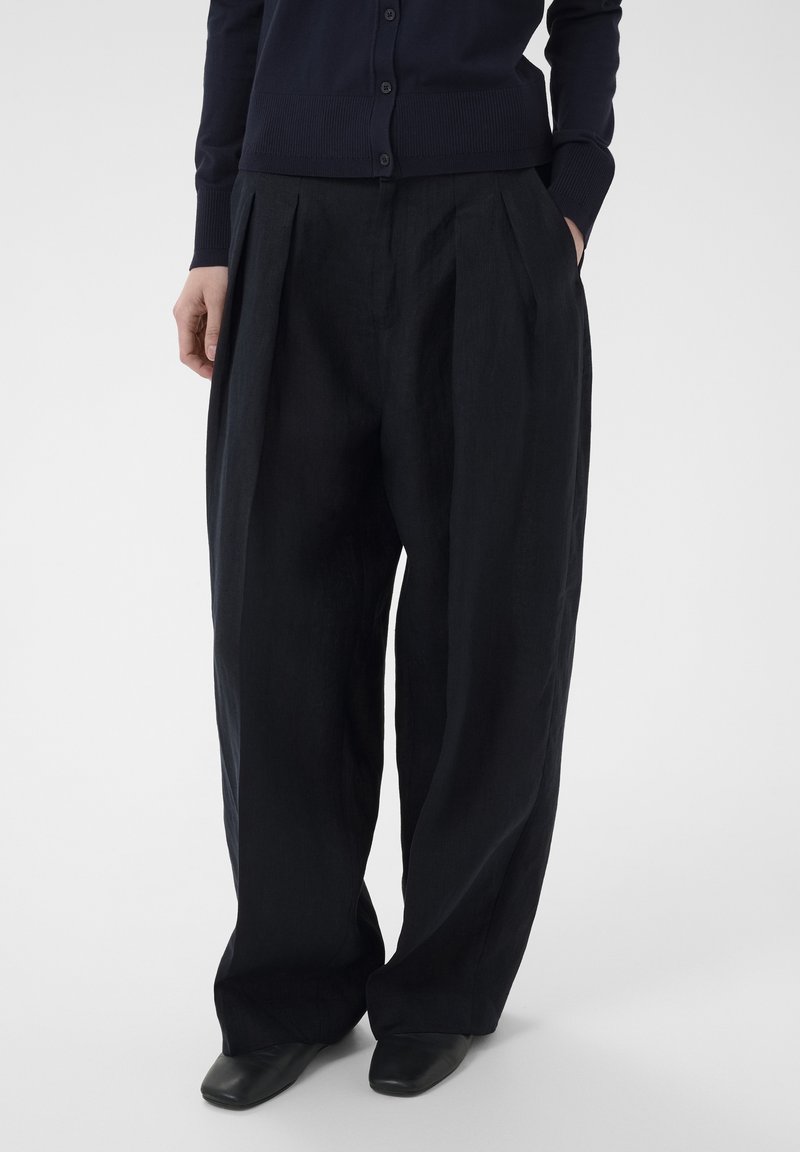 Personne portant un pantalon large noir à plis avant, un cardigan noir boutonné, les mains dans les poches, et des chaussures noires à bout carré.