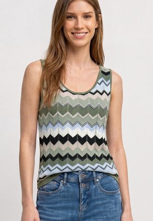 Młoda kobieta uśmiechająca się, ubrana w bezrękawnik z dzianiny z zielono-czarno-niebieskim wzorem w zygzak oraz niebieskie dżinsy z podwójnym zapięciem na guziki.