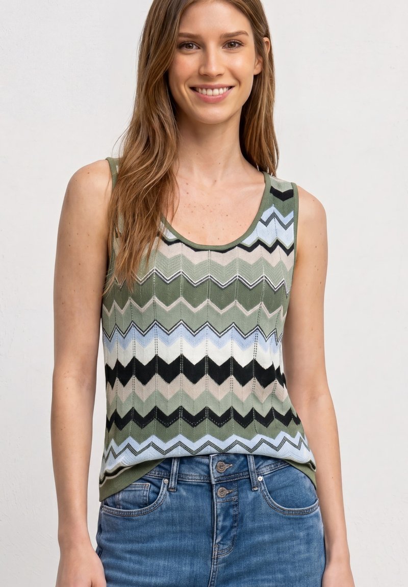 Jeune femme souriante, portant un haut en maille sans manches avec un motif zigzag vert, noir et bleu, et un jean en denim bleu avec une fermeture à double bouton.