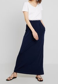 Námořnická modrá maxi sukně vyrobená z měkké, elastické tkaniny s bočními kapsami. Nosit s bílým tričkem s výstřihem do V a černými plochými sandály.