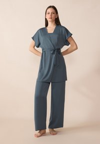 Ľahký loungewear set v tmavom teal odtieni so zavinovacou blúzkou s opaskom a širokými rukávmi, doplnený o nohavice s širokými nohavicami. Hladká textúra.