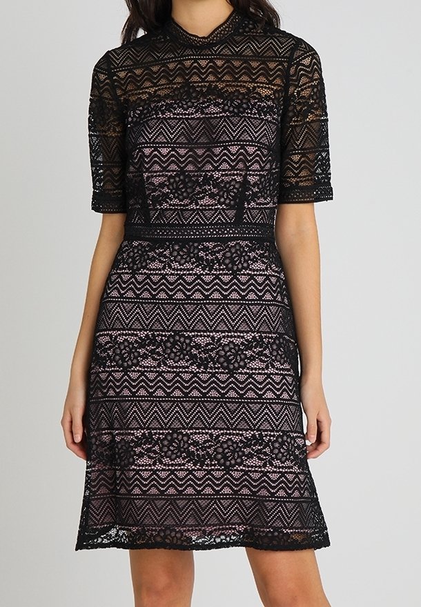 Femme portant une robe en dentelle noire mi-longue aux genoux avec des motifs géométriques et floraux sur une doublure rose clair, dotée de manches courtes et d'un col montant.