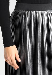 Falda plisada en negro y plata con un acabado brillante texturizado, que presenta una cinturilla ancha negra. La mano descansa ligeramente sobre la tela.