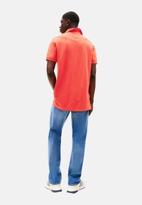 Camisa polo de coral brillante con mangas cortas, cuello acanalado y un corte relajado, combinada con unos jeans de mezclilla azul claro y zapatillas blancas.