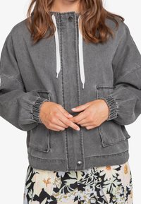 Chaqueta de mezclilla gris con un ajuste relajado, puños elásticos, cordones blancos, bolsillos frontales y cierre de botón a presión. Sin patrones visibles.