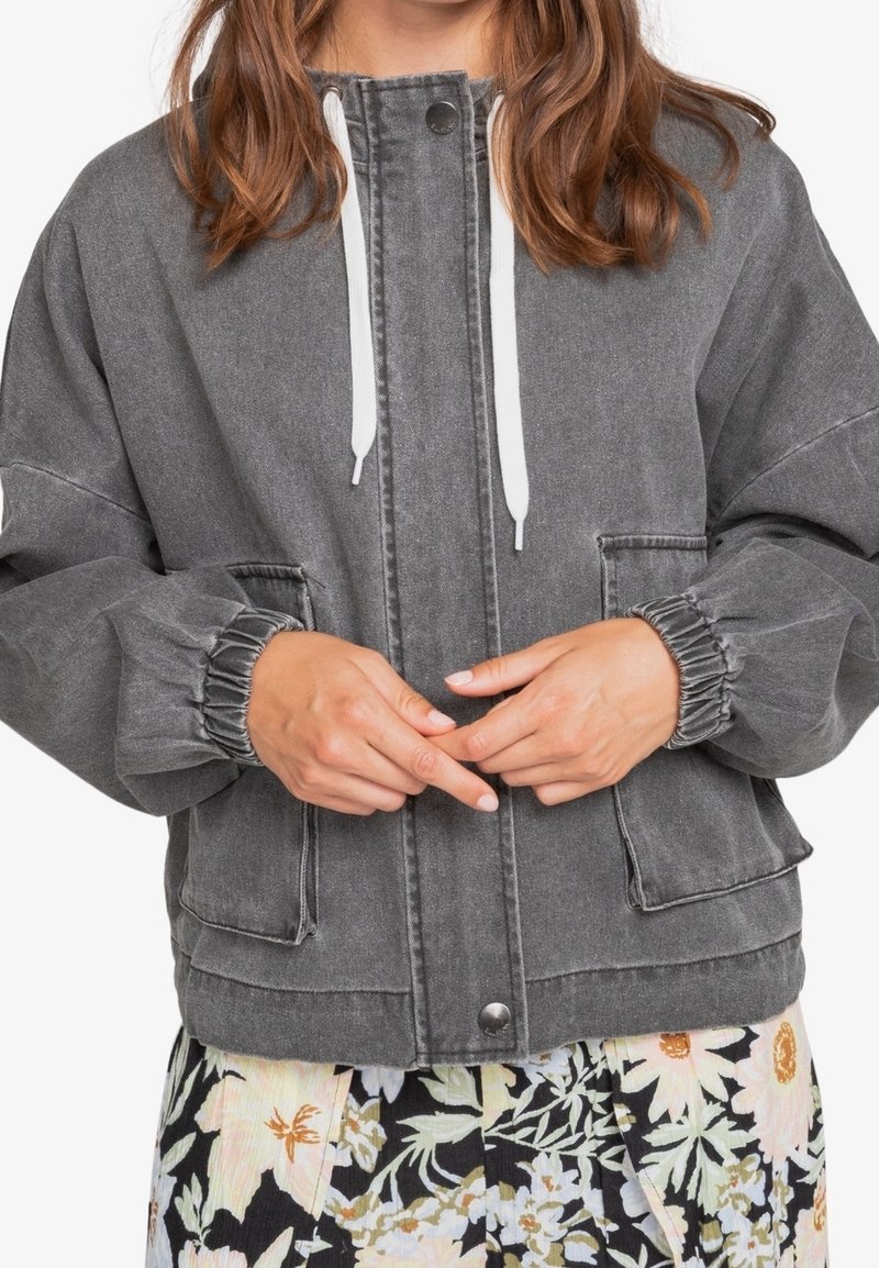 Chaqueta de mezclilla gris con un ajuste relajado, puños elásticos, cordones blancos, bolsillos frontales y cierre de botón a presión. Sin patrones visibles.