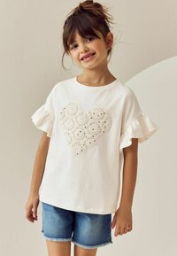 Weißes Baumwoll-T-Shirt mit Rüschenärmeln, versehen mit einer strukturierten Herz-Applikation aus Spitze und Pailletten. Kombiniert mit blauen Jeansshorts.