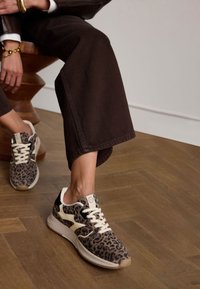 Leopardenmuster-Sneaker mit cremefarbenen Akzenten, strukturiertem Stoff, weißen Schnürsenkeln und einer gesprenkelten Sohle, kombiniert mit braunen, weiten Hosen.