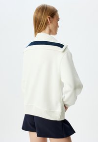 Sudadera blanca con cuello de canalé y rayas marinas, que presenta puños y dobladillo de canalé. La parte posterior tiene un corte recto y un diseño de ajuste relajado.