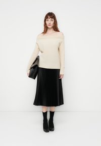 Suéter de malha bege com um ombro à mostra emparelhado com uma saia midi plissada preta. A modelo segura uma mala preta, vestindo meias sociais pretas e saltos.