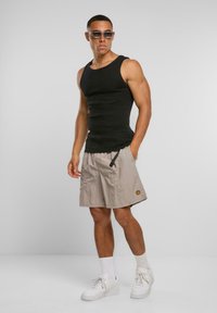 Schwarzes geripptes Tanktop, beige Shorts mit einem Smiley-Patch, weiße Turnschuhe und klare Sonnenbrille. Das Model steht vor einem neutralen Hintergrund.