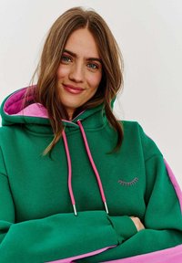 Zelená mikina s ružovými akcentmi, s kapucňou na šnúrku, vyšívanými detailmi rias a vo voľnom strihu. Mäkko texturovaná tkanina.