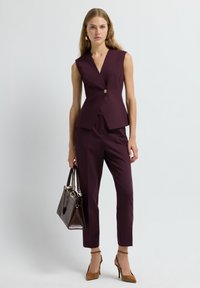 Vrouw in een mouwloze paarse blazer en tapse broek, met een bruine handtas en beige hoge hakken tegen een effen achtergrond.