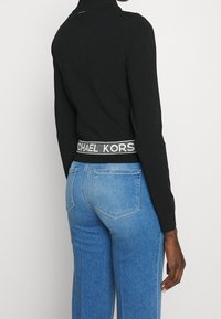 Maglione a collo alto nero con un design aderente, abbinato a jeans in denim blu chiaro. La vita presenta un evidente dettaglio con logo bianco.
