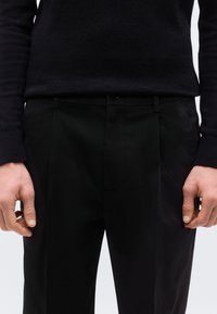 Μαύρο παντελόνι με λεία υφή, διαθέτει μέση με κούμπωμα με κουμπί, επίπεδη μπροστινή όψη και διακριτικές πιέτες για επιπλέον λεπτομέρεια.