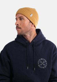 Bonnet côtelé jaune fabriqué en matériau doux, porté avec un sweat à capuche bleu marine portant un logo circulaire sur la poitrine.