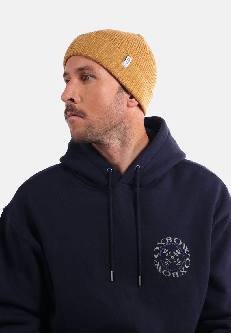 Bonnet côtelé jaune fabriqué en matériau doux, porté avec un sweat à capuche bleu marine portant un logo circulaire sur la poitrine.