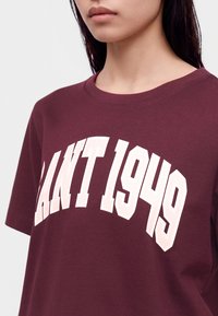 Νέος άνθρωπος φοράει ένα βυσσινί μπλουζάκι με μεγάλα λευκά γράμματα που γράφουν «"GANT 1949"» στο στήθος.
