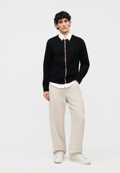 Jeune homme portant un cardigan noir avec une bande colorée sur une chemise blanche, un pantalon large beige et des baskets blanches, debout devant un fond blanc.