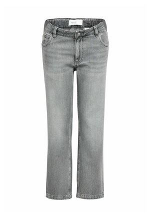 Gravid kvinde går fremad iført lys beige langærmet skjorte, grå jeans og lyserøde flettede sko mod en ensfarvet brun baggrund.